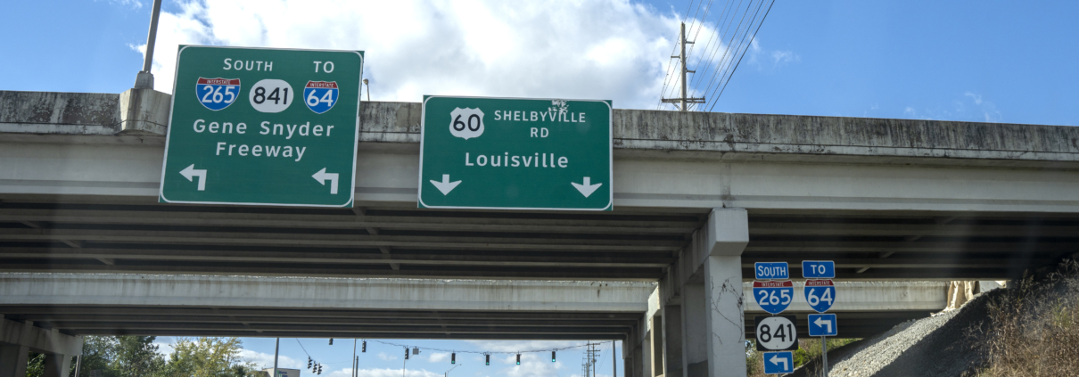 Two traffic signs attached to the Gene Snyder Freeway overpass over Shelbyville Road. The signs show left turning traffic will enter the Gene Snyder. Straight traffic will continue on to Louisville.