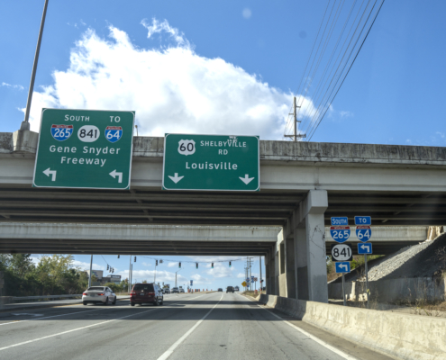 Two traffic signs attached to the Gene Snyder Freeway overpass over Shelbyville Road. The signs show left turning traffic will enter the Gene Snyder. Straight traffic will continue on to Louisville.